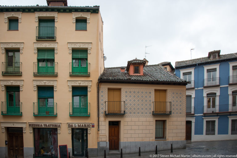 Ochre, patterned beige, and blue façades with balconies, green shutters, tiled roofs, and ground-floor shops.
