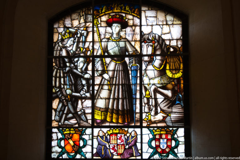 Stained-glass window with a standing robed figure in a red cap, mounted attendants, and three heraldic shields along the bottom.
