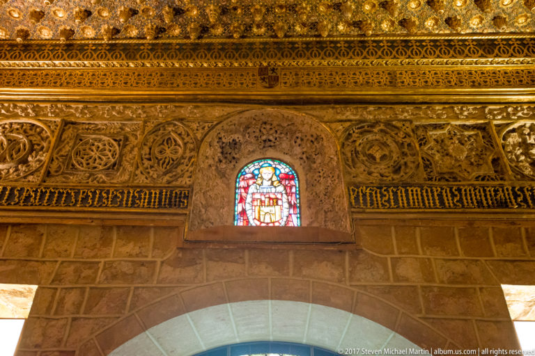 Stone arch topped by a small heraldic stained-glass opening and a dense gilded carved frieze of foliage and medallions.