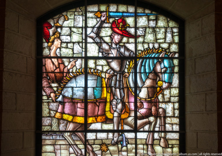 Stained-glass window showing an armored rider on a horse with blue trappings, surrounded by several smaller attendant figures.