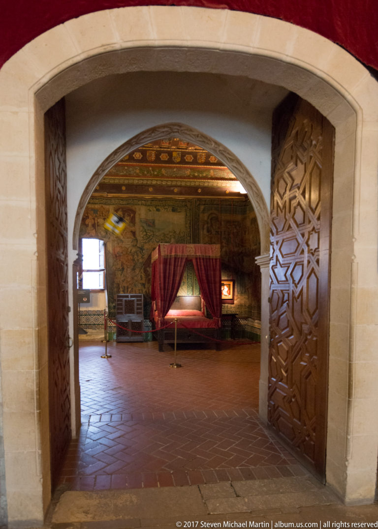 View through carved wooden doors and a broad arch into a decorated chamber with a curtained bed and warm-toned ceiling.