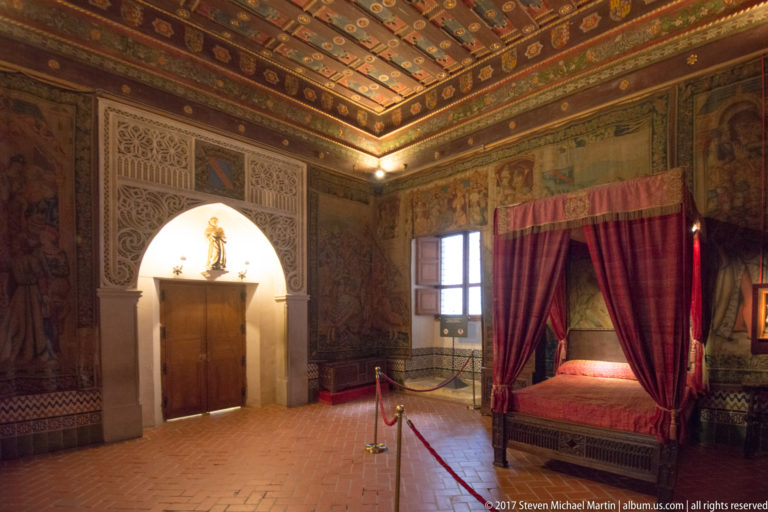 Historic chamber with a red-curtained bed, painted wall decoration, patterned panels, timber ceiling, and an arched doorway.