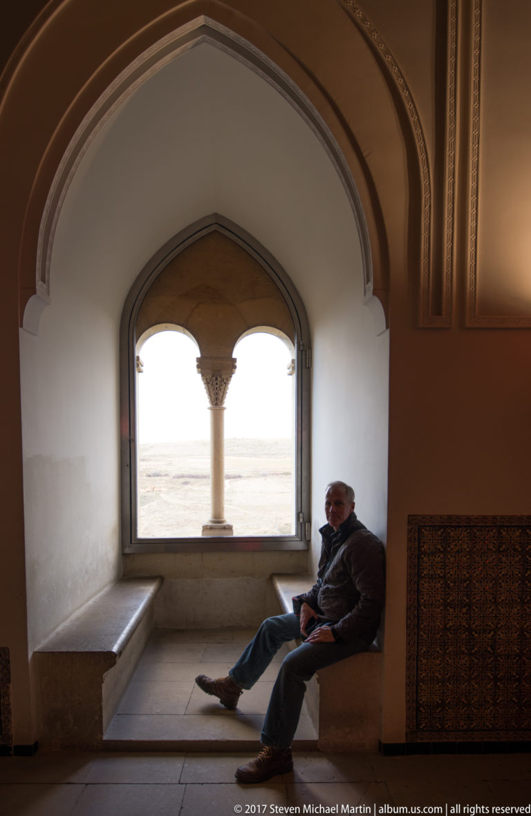 Seated visitor in a deep stone window alcove beside a twin-arched opening divided by a slender central column.