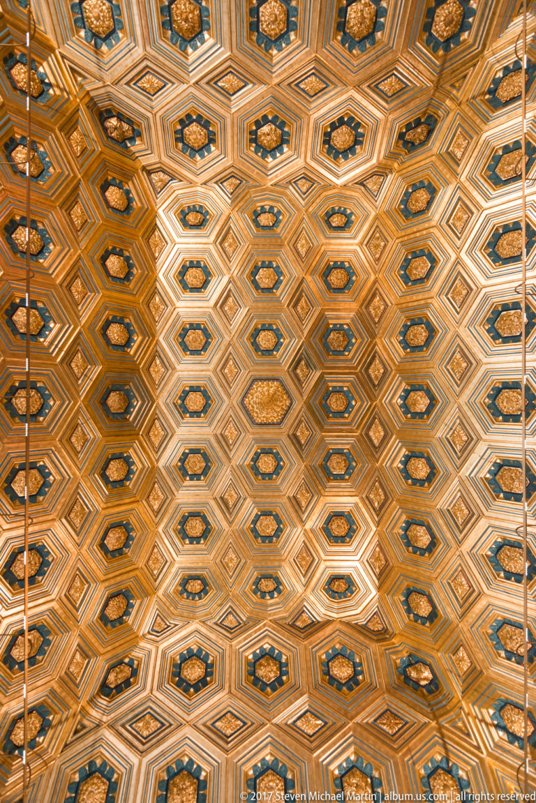 Close upward view of a coffered ceiling patterned with interlocking hexagons and diamonds in gold, blue, white, and wood tones.