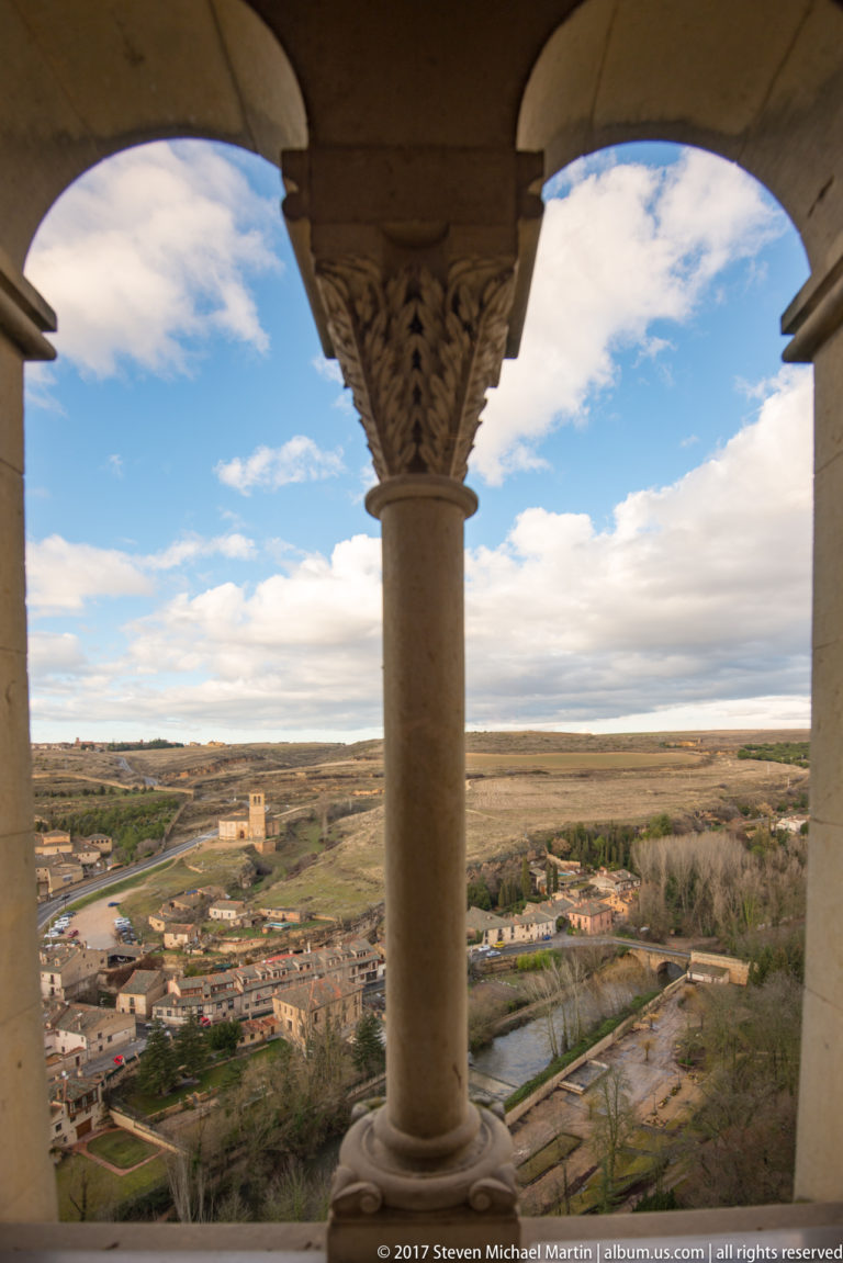 Carved central column between two arches frames a view over low buildings, a river corridor, and the open plateau beyond Segovia.