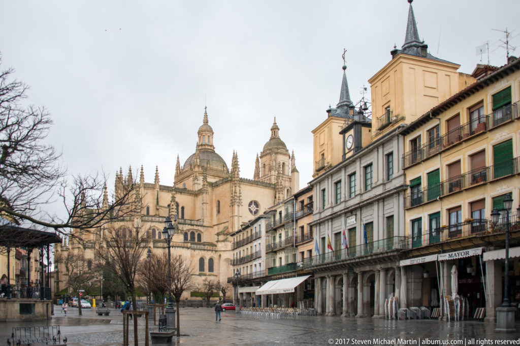 Segovia Cathedral with pinnacles, dome, and tower rising behind arcaded buildings and wet paving in Plaza Mayor.