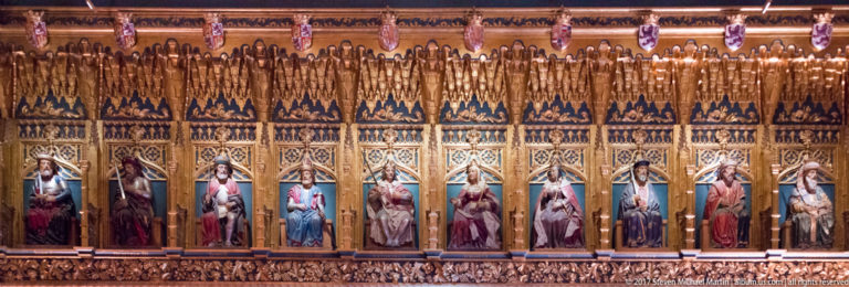Long row of seated polychrome royal figures beneath a continuous gilded carved canopy, with heraldic shields repeating above.