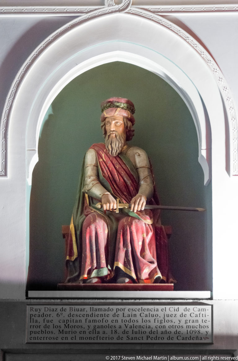 Bearded seated ruler in a red robe holds a long horizontal staff inside a white arched niche with Spanish text panels.