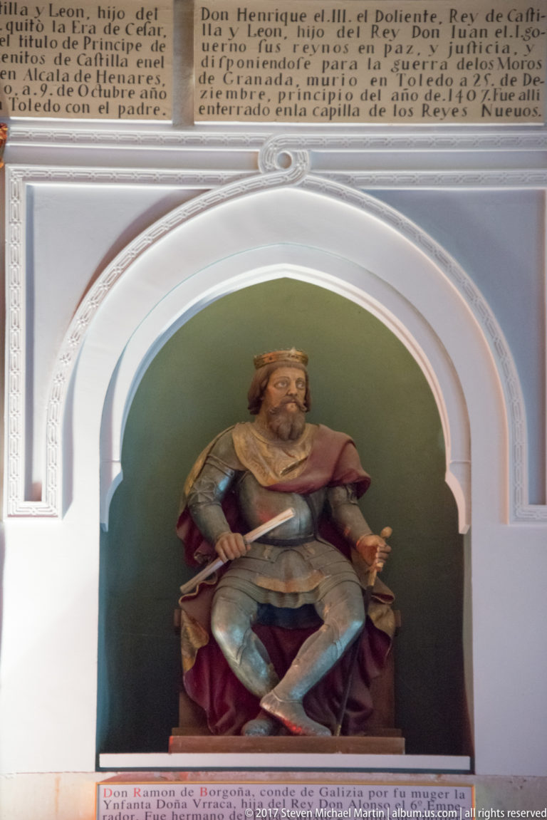 Seated crowned ruler in blue and red garments inside a white arched niche, with a long weapon and Spanish inscription below.