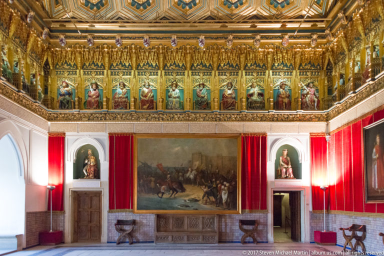 Historic palace room with a continuous upper gallery of royal figures, red drapery, wall paintings, carved furnishings, and side niches.