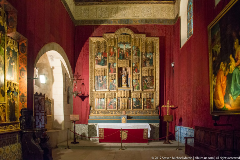 Small red-walled chapel with a tall multi-panel altarpiece above a stone altar, flanked by paintings and dark wooden furnishings.