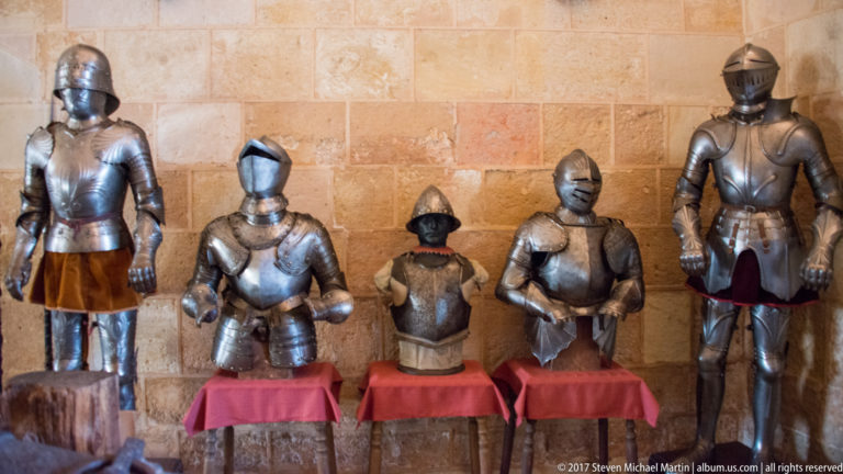 Two tall suits of plate armor flank several smaller torso and helmet displays on red-draped stands against a stone wall.
