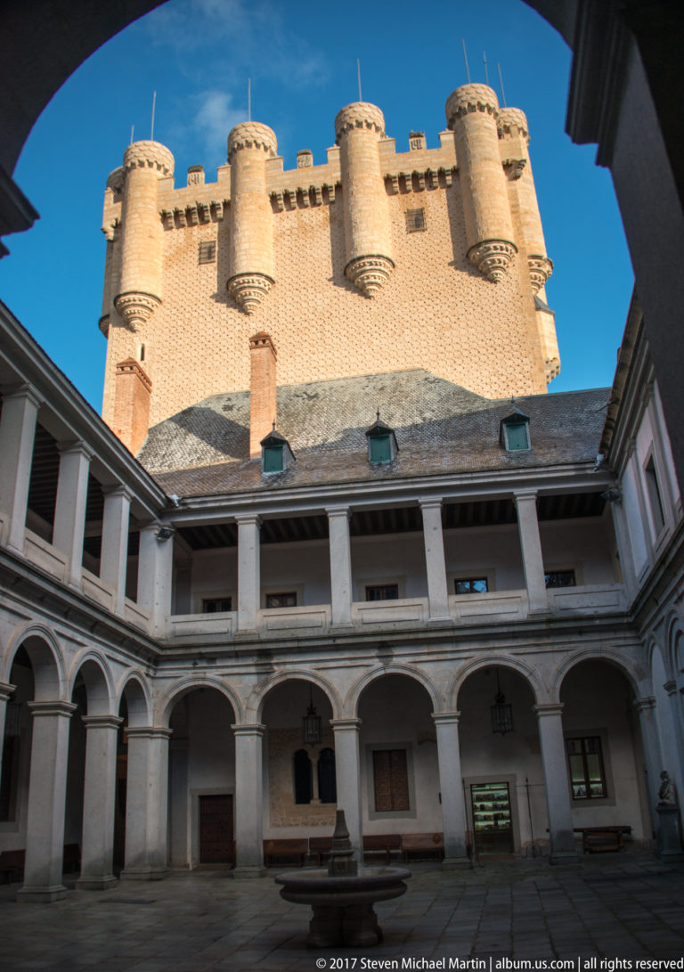 Arcaded two-story courtyard beneath a massive crenellated brick tower with projecting cylindrical corner turrets against a blue sky.