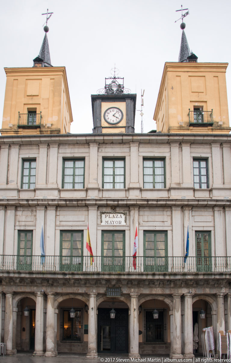 Symmetrical civic façade on Plaza Mayor with stone arcade, central balcony, flags, rooftop clock, and two end pavilions.