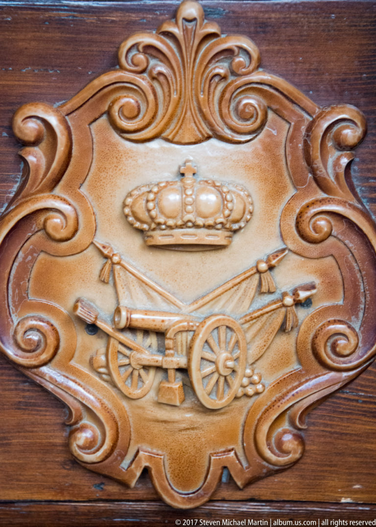 Carved wooden plaque with a crown above crossed artillery pieces and a wheeled cannon inside a scrolling ornamental border.