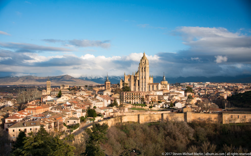 Elevated view across Segovia with the cathedral tower rising above red-tiled roofs and distant mountains under layered clouds.