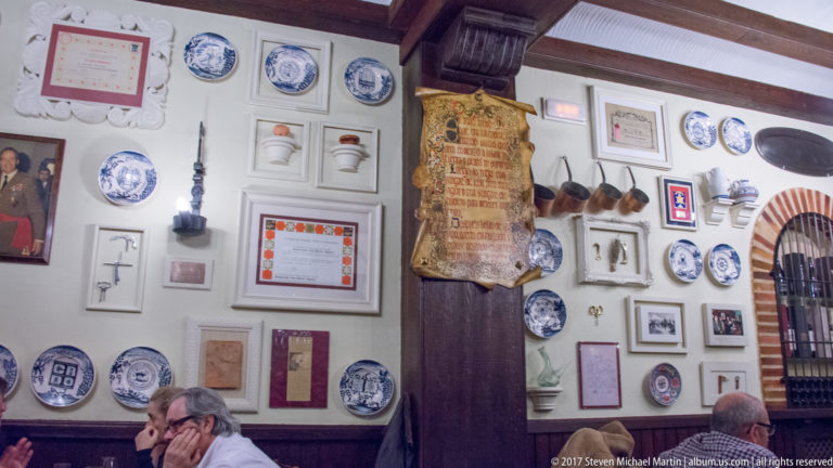 Restaurant wall densely covered with blue-and-white plates, framed documents, utensils, and memorabilia above seated diners.