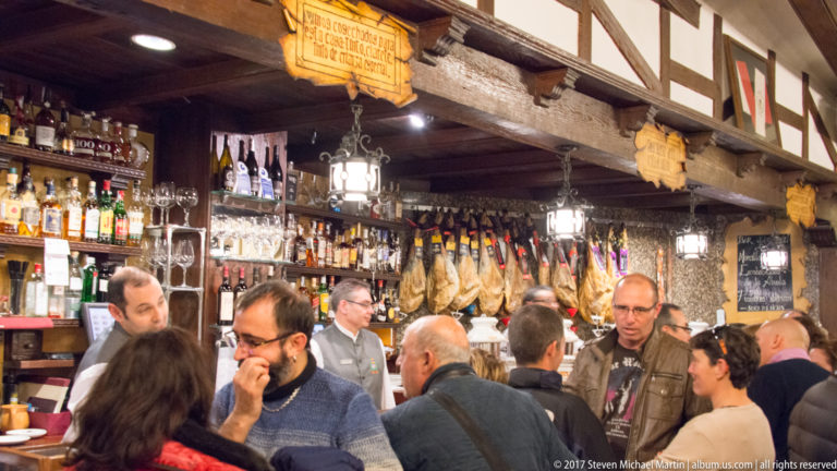 Busy restaurant with diners and staff beneath timber beams, hanging lanterns, shelves of bottles, and rows of cured hams.
