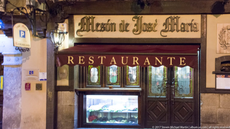 Entrance to Mesón de José María with a large restaurant sign, lit display case, dark paneled doors, and stone wall surround.