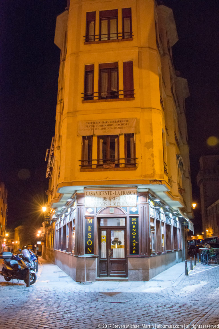 Night view of a multi-story corner building with a brightly lit ground-floor restaurant and shuttered upper windows.