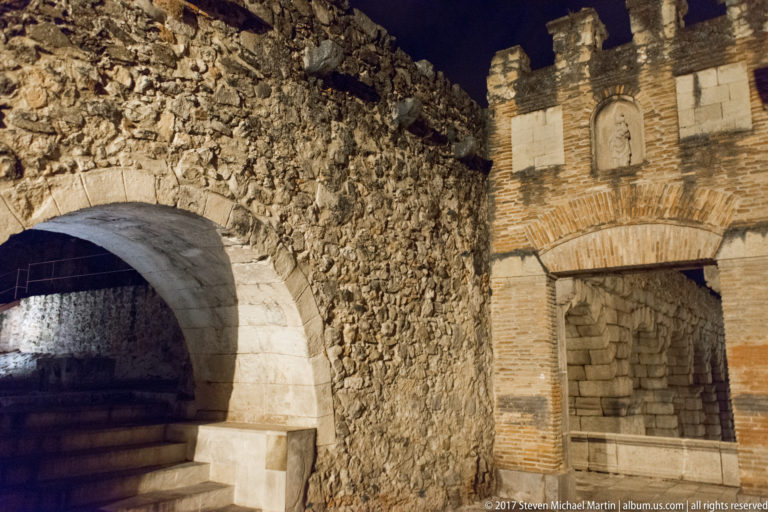 Night view of a broad stone arch beside a taller brick-and-stone wall with a recessed niche and crenellated top.