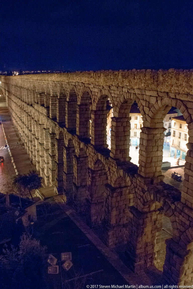 Segovia’s Roman aqueduct runs diagonally through the city at night, with two tiers of granite arches illuminated against a dark blue sky.