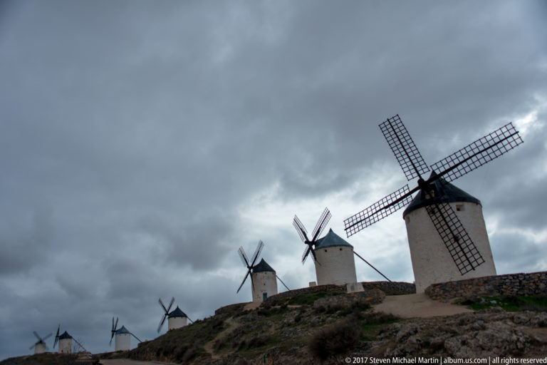 Line of white tower windmills with lattice sails extending across a rocky ridge beneath dark storm clouds.