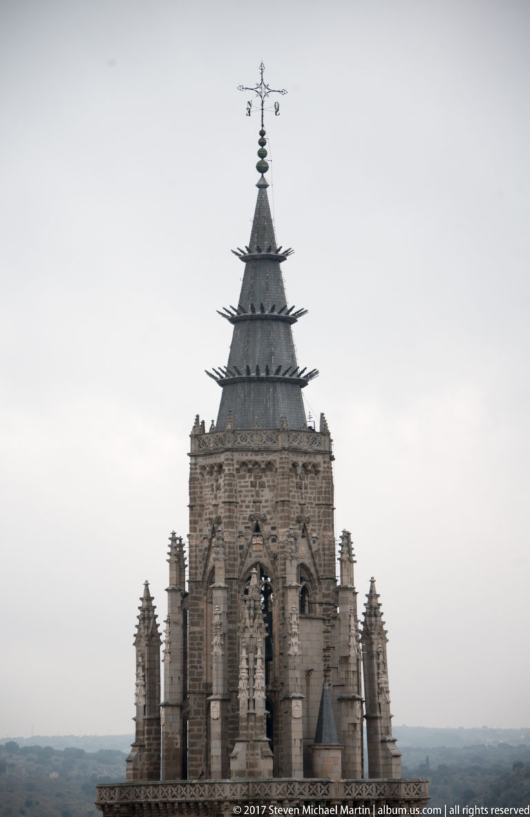 Close telephoto view of Toledo Cathedral tower with Gothic tracery, pinnacles, sculpted details, and a steep metal-clad spire.