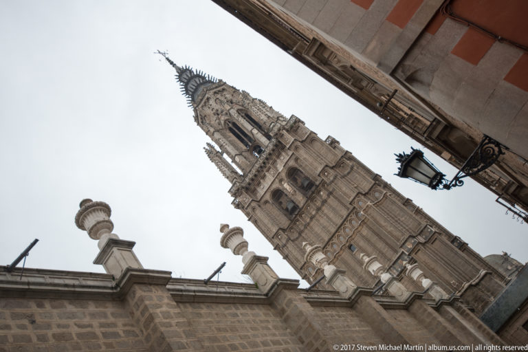 Low-angle view of Toledo Cathedral tower rising between stone façades, decorative finials, and a hanging street lantern.