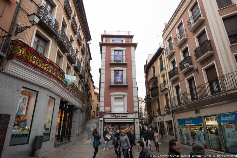 Busy Toledo pedestrian street with shops, balconies, narrow multistory buildings, and a tall slender red façade at center.
