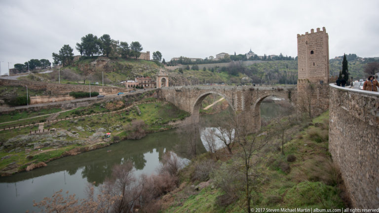 Historic stone bridge with multiple arches and a square defensive tower crossing the Tagus River below Toledo’s walls.