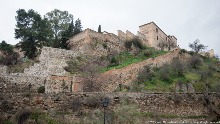 Stone defensive walls and terraces climbing a steep green rocky hillside beneath historic buildings in Toledo.