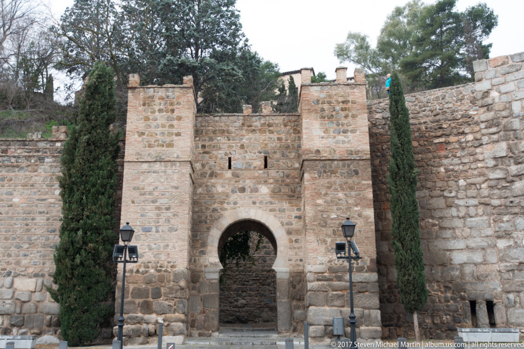 Small round-arched gateway set in thick Toledo stone walls, flanked by square masonry and tall cypress trees.