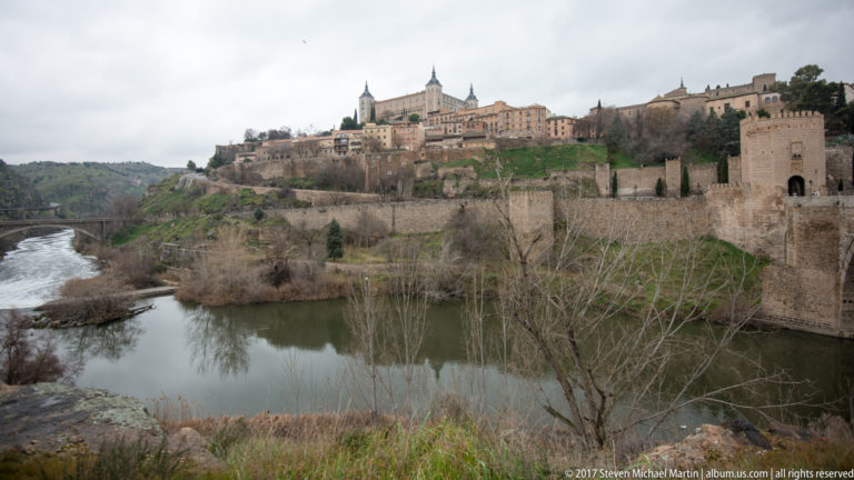 Toledo’s walls and historic skyline rising above a bend of the Tagus River with winter trees along the bank.