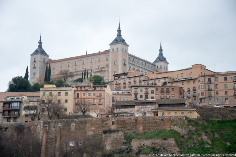 Alcázar of Toledo with four corner towers rising above houses, walls, and terraced slopes of the historic city.