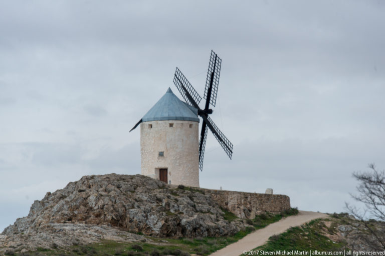 White tower windmill with crossed sails standing on a rocky outcrop above a curving path and low stone wall.