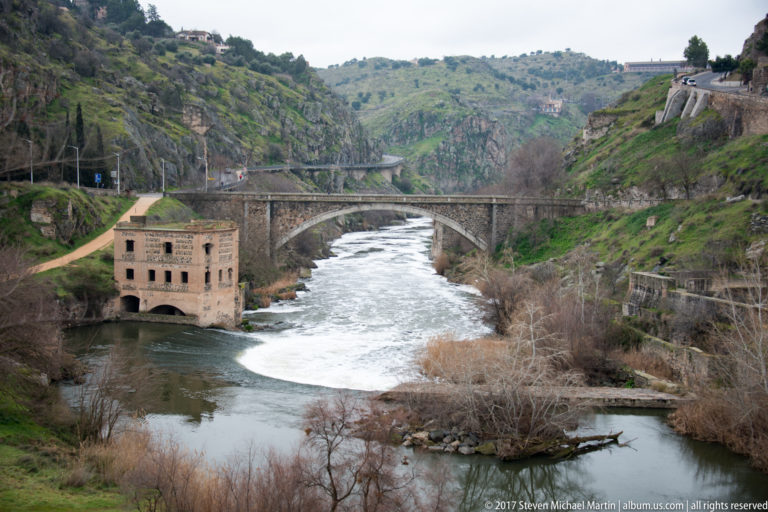 Stone bridge over the Tagus beside a historic riverside building, with fast water, rocky slopes, and another bridge upstream.