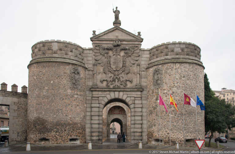 Puerta de Bisagra with central arched entrance, twin round towers, imperial eagle relief, and flags beside the gateway.