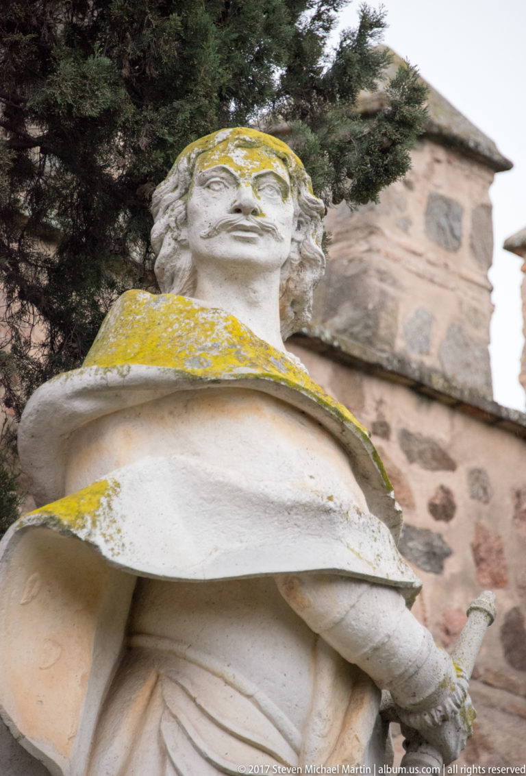 Low-angle close view of a weathered robed stone statue holding a narrow object, with yellow staining and masonry behind.
