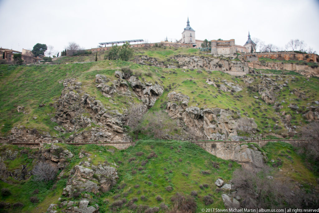 Rocky green gorge beneath Toledo with historic buildings and towers along the ridge under an overcast sky.