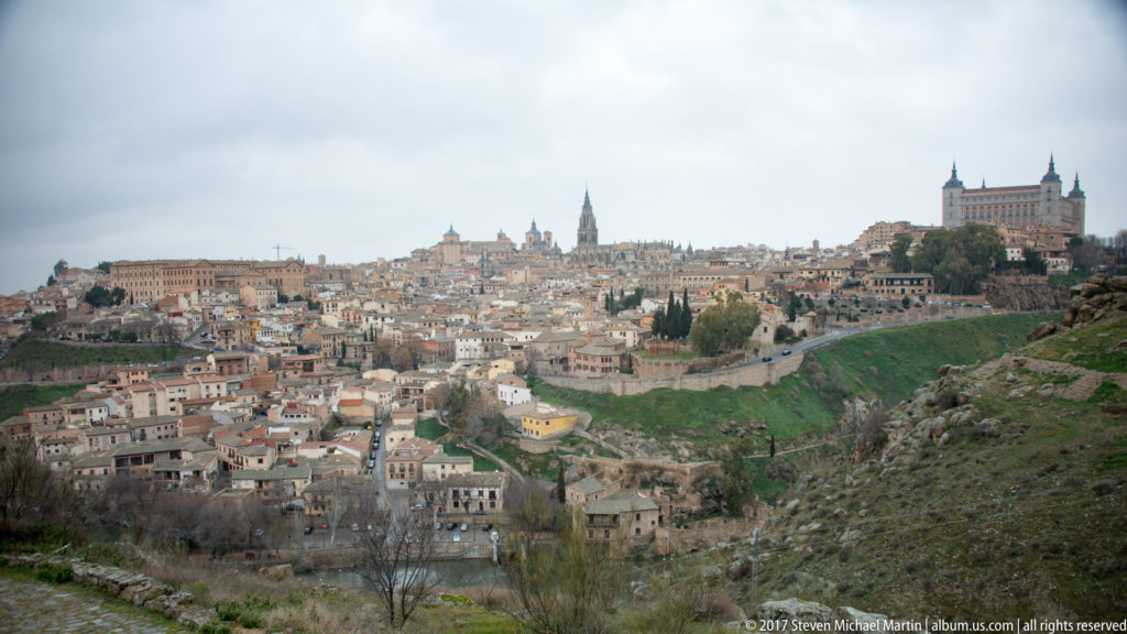 Wide Toledo panorama with dense historic buildings, cathedral spire, Alcázar, walls, and green slopes above the gorge.