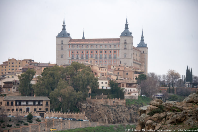 Telephoto view of the Alcázar of Toledo with four corner towers above houses, trees, rock, and defensive walls.