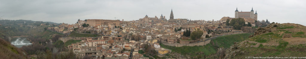 Distant panoramic view of Toledo with clustered historic buildings, cathedral tower, Alcázar, walls, and the Tagus gorge below.