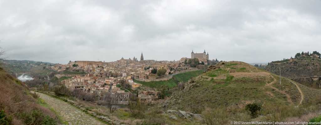 Wide view from a hillside path toward Toledo’s skyline, with the cathedral, Alcázar, open slopes, and deep gorge beneath gray clouds.