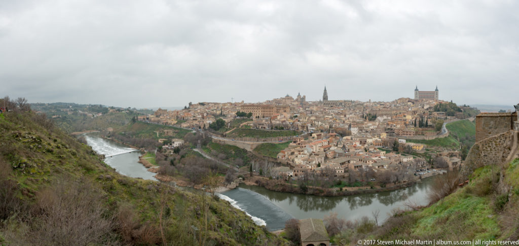 Wide panorama of Toledo on a hill encircled by the Tagus River, with cathedral and Alcázar visible beneath gray clouds.