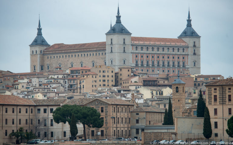 Telephoto view of the Alcázar of Toledo with four corner towers rising above clustered historic buildings.