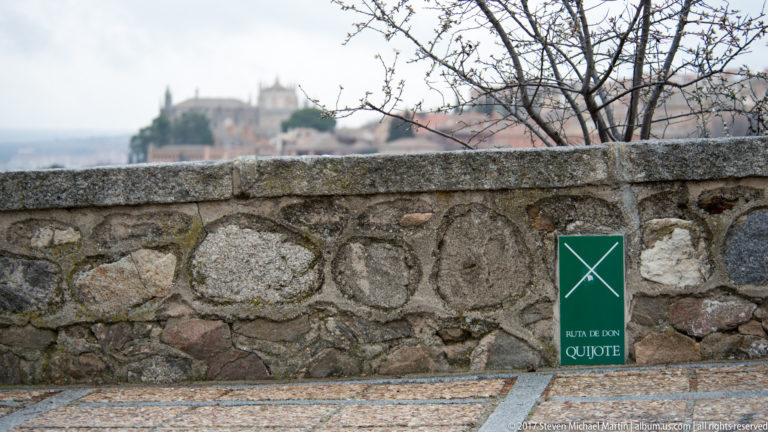 Green Ruta de Don Quijote marker beside a rough stone wall, with Toledo blurred in the distance.