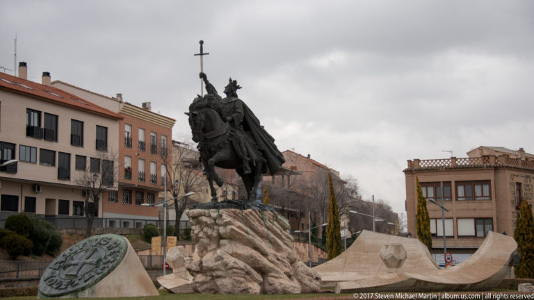 Bronze equestrian statue of a medieval ruler raising a sword above a rugged stone base in a Toledo plaza.