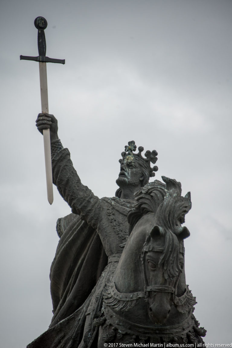 Low-angle close view of a bronze medieval rider raising a sword while mounted on a strongly modeled horse.