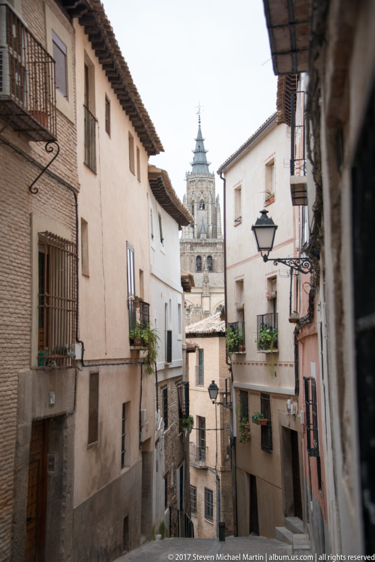 Narrow Toledo street lined with stucco and brick buildings, balconies, lamps, and an ornate church tower in the distance.